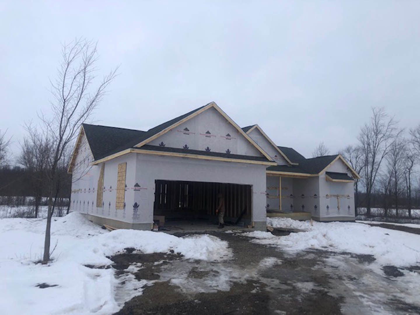 Framed house under construction with exposed wood, snow covering ground, man standing in open garage, leafless trees in background, overcast winter sky