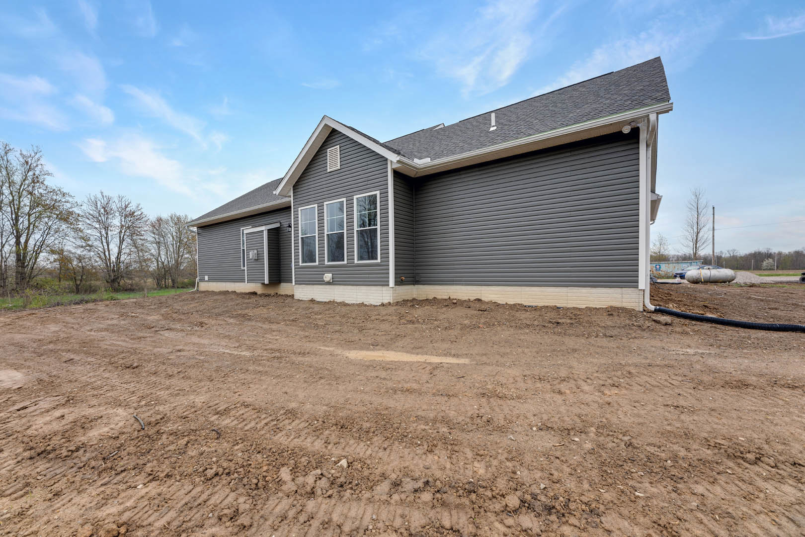 Partially built house with exposed wooden framing, white window frames, bare dirt yard, leafless tree, and cloudy sky in the background