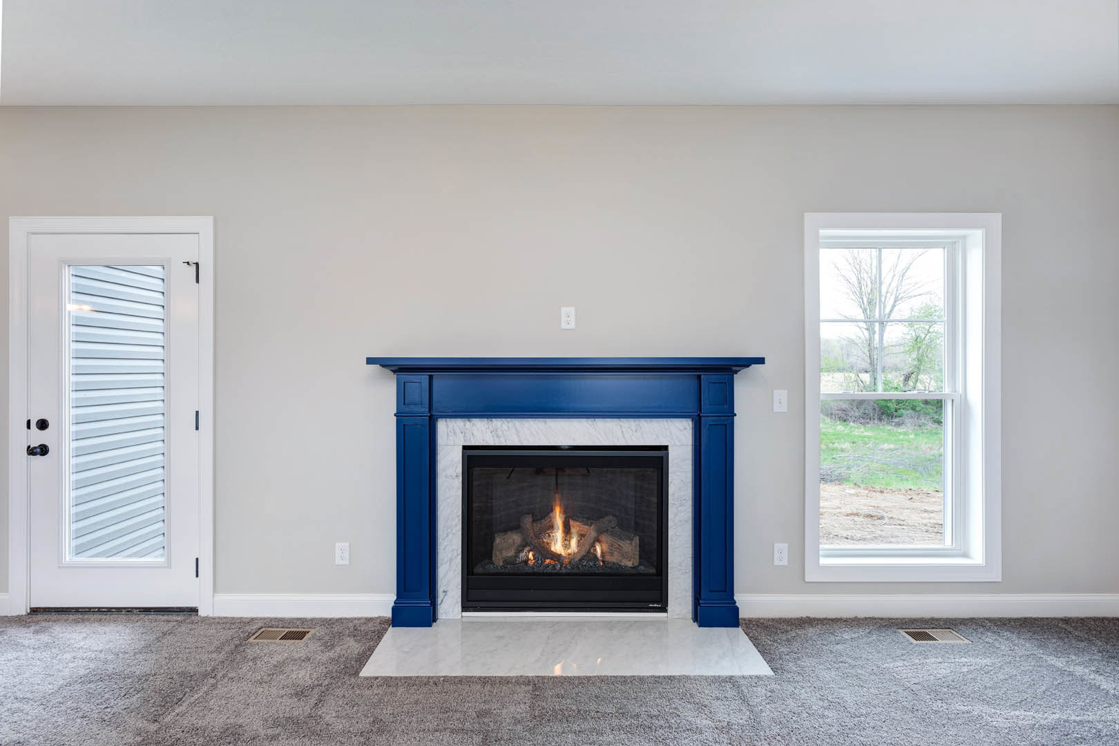 Blue-trimmed fireplace with burning fire, white door with black handle, window overlooking field and trees, white surface with black border, hardwood floor