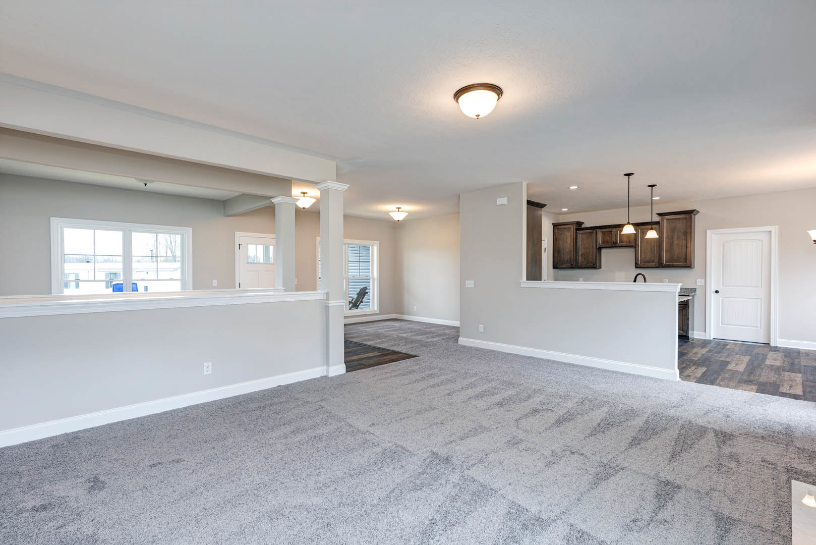 Open concept living room with carpet flooring, adjacent kitchen and dining area, white door with black handle, ceiling light fixture, large window, person standing near window