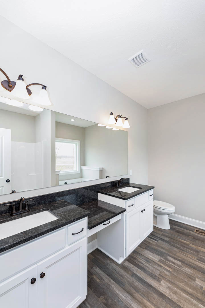 Bathroom with a wide mirror above a white sink, white toilet with seat down, three-light fixture mounted above mirror, window overlooking grassy field, ceiling vent, light-colored