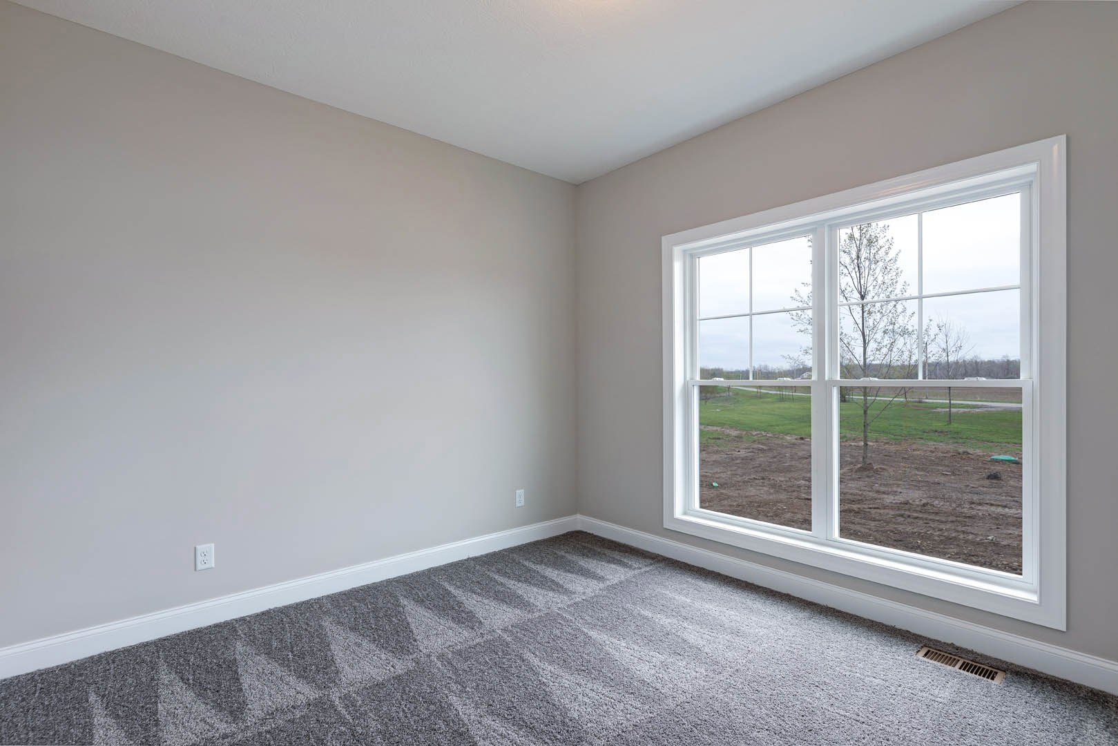 Bedroom with grey carpet, white walls, large window overlooking grassy field and tree, white ceiling, floor vent visible near window