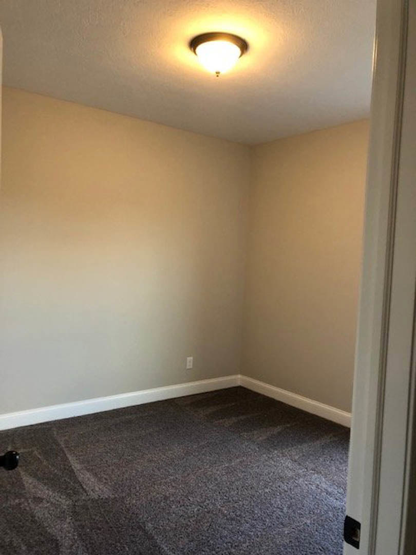Carpeted room with white plaster walls, ceiling molding, and a modern ceiling light fixture; partial view of a door and laminate flooring.