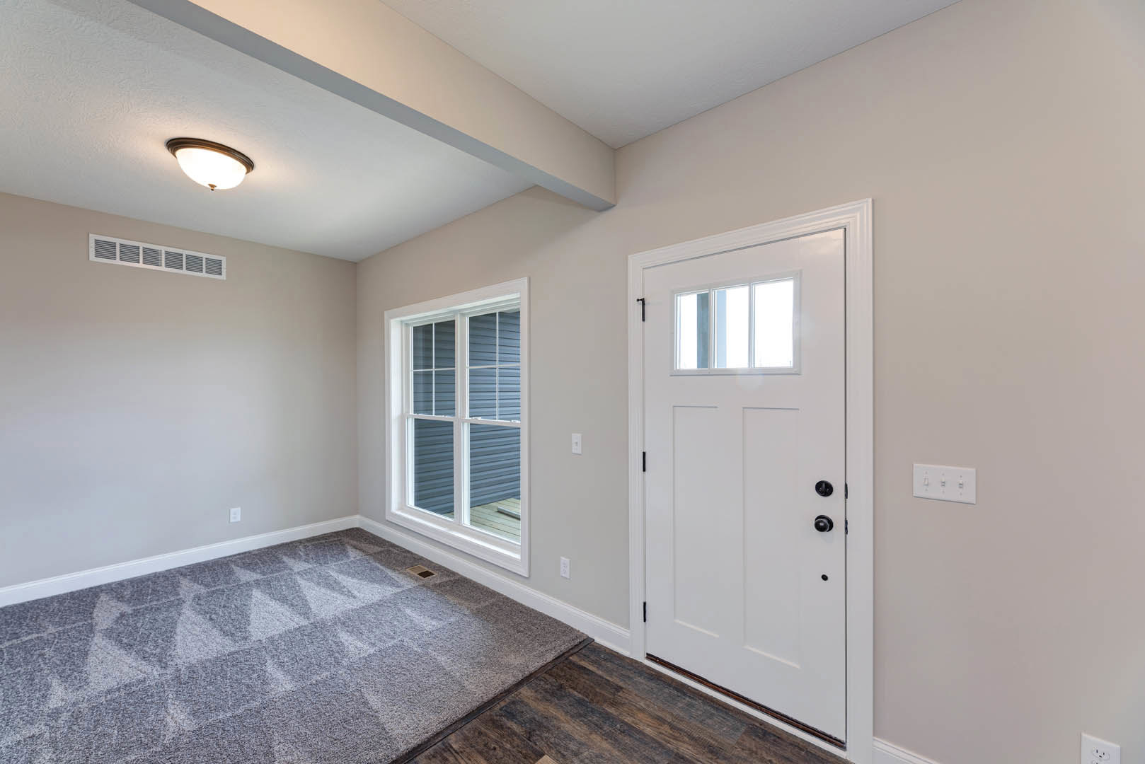 White paneled door with black hardware, large window with white trim, grey carpet featuring triangle pattern, white walls, ceiling-mounted light fixture, and nearby electrical