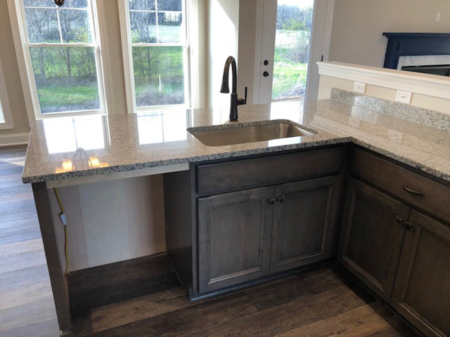 Modern kitchen featuring a black faucet and stainless steel sink set into a light stone countertop, white tile backsplash, wood cabinetry, and a window overlooking green trees.