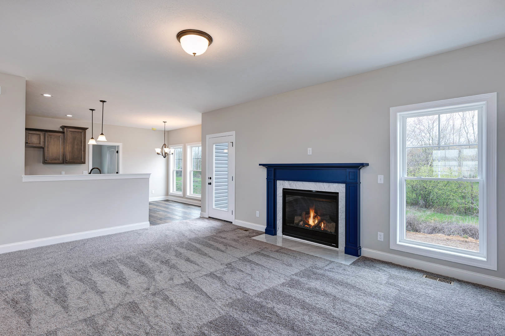 Open concept living room with carpeted floor, stone fireplace with active fire, adjacent kitchen, large window overlooking tree-filled field, white door with black hardware, modern