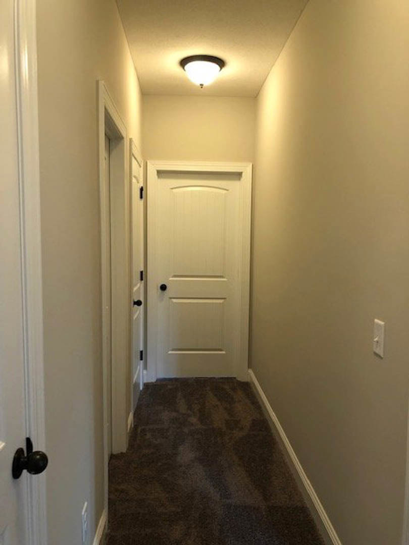 Hallway with white walls, dark carpeted staircase, white door featuring black knobs, ceiling-mounted light fixture, and light switch on the wall