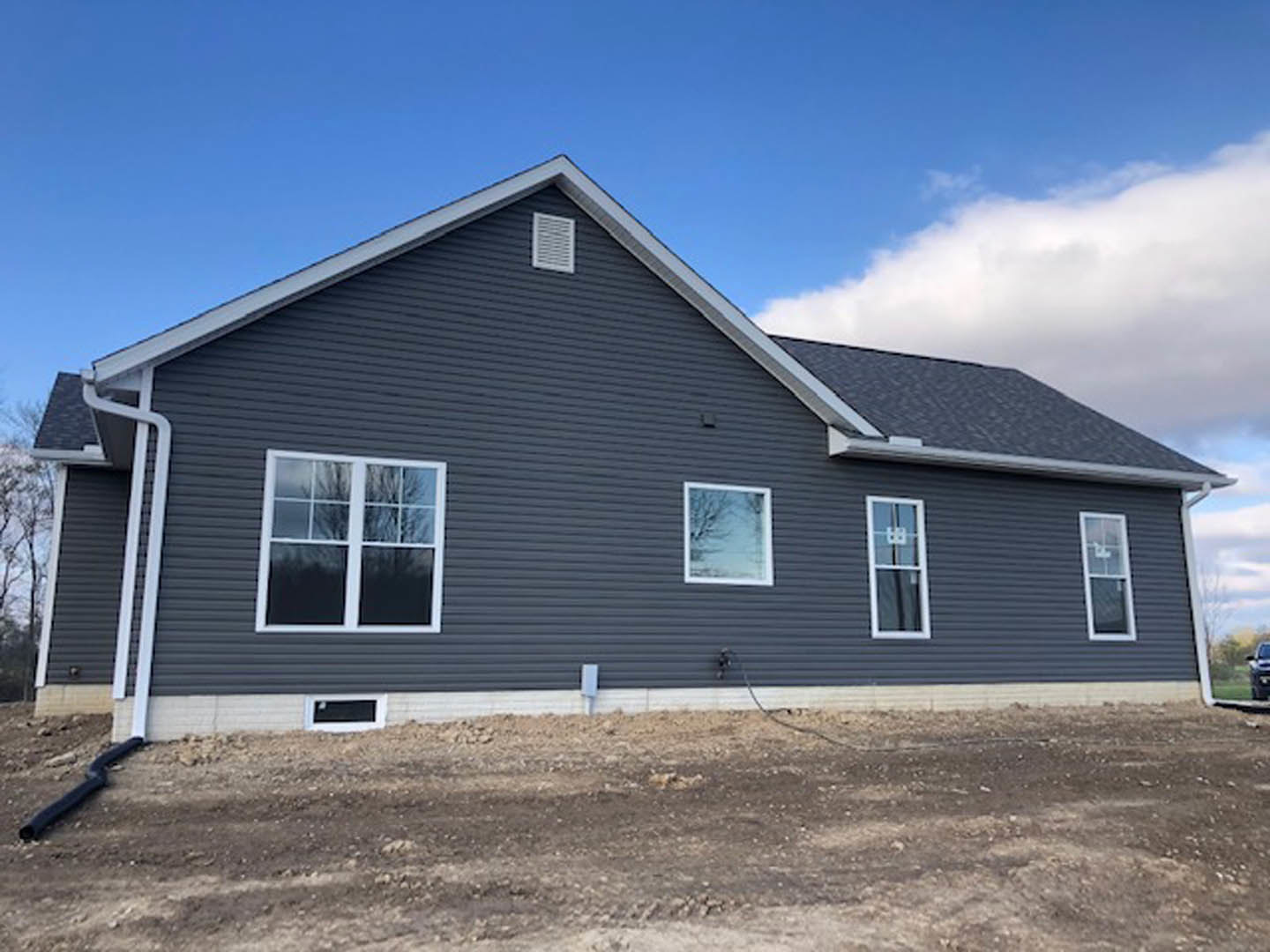 Modern house with black siding, white-framed windows, white vent, and dirt yard featuring a metal pole; blue sky and tree visible in background