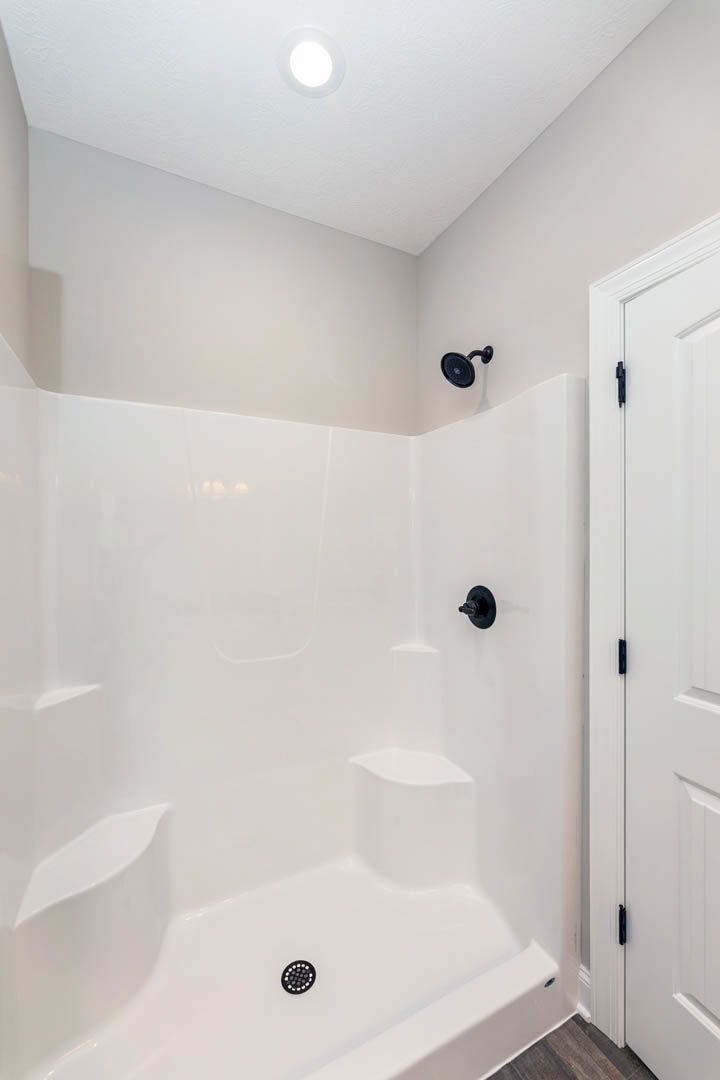White tiled shower with black showerhead and handle, white door with black hardware, circular ceiling light, and smooth white walls.