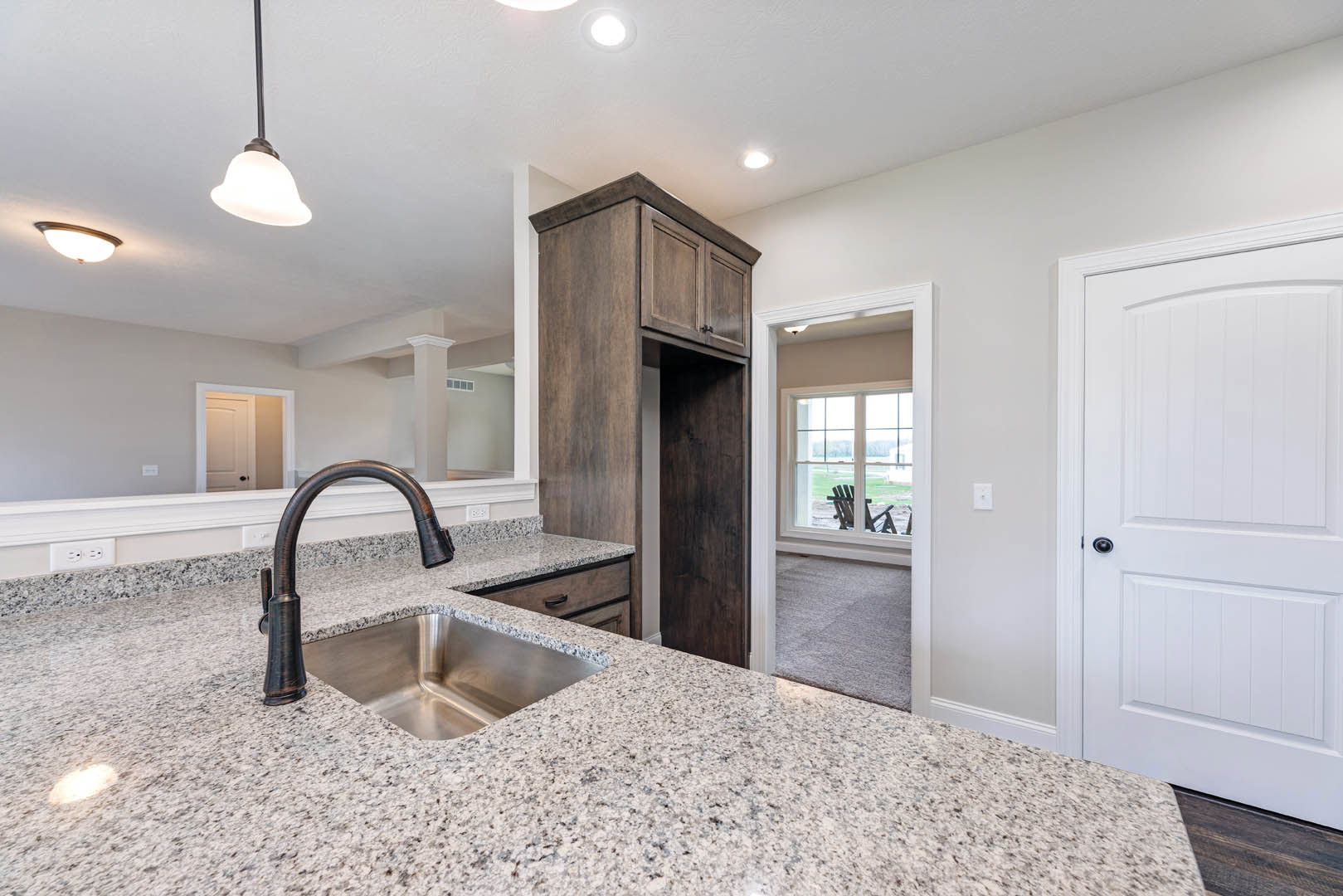 Kitchen featuring a stainless steel sink set in a granite countertop, wood cabinetry with modern hardware, white tile backsplash, black-handled white door, chair positioned by a