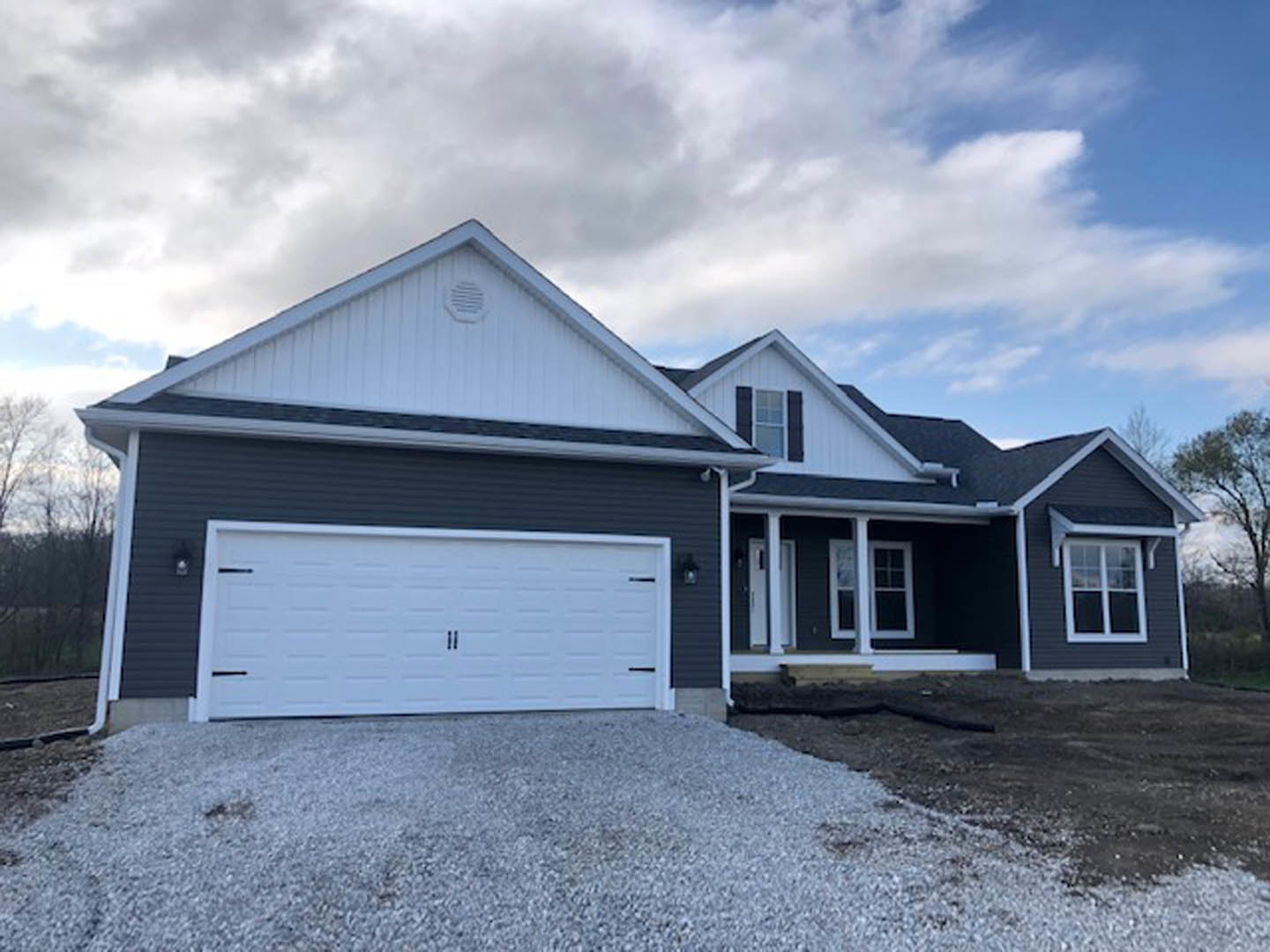 White garage door with black trim, grey gravel driveway, white-framed window, light-colored siding, wall vent, cloudy sky, and trees in background