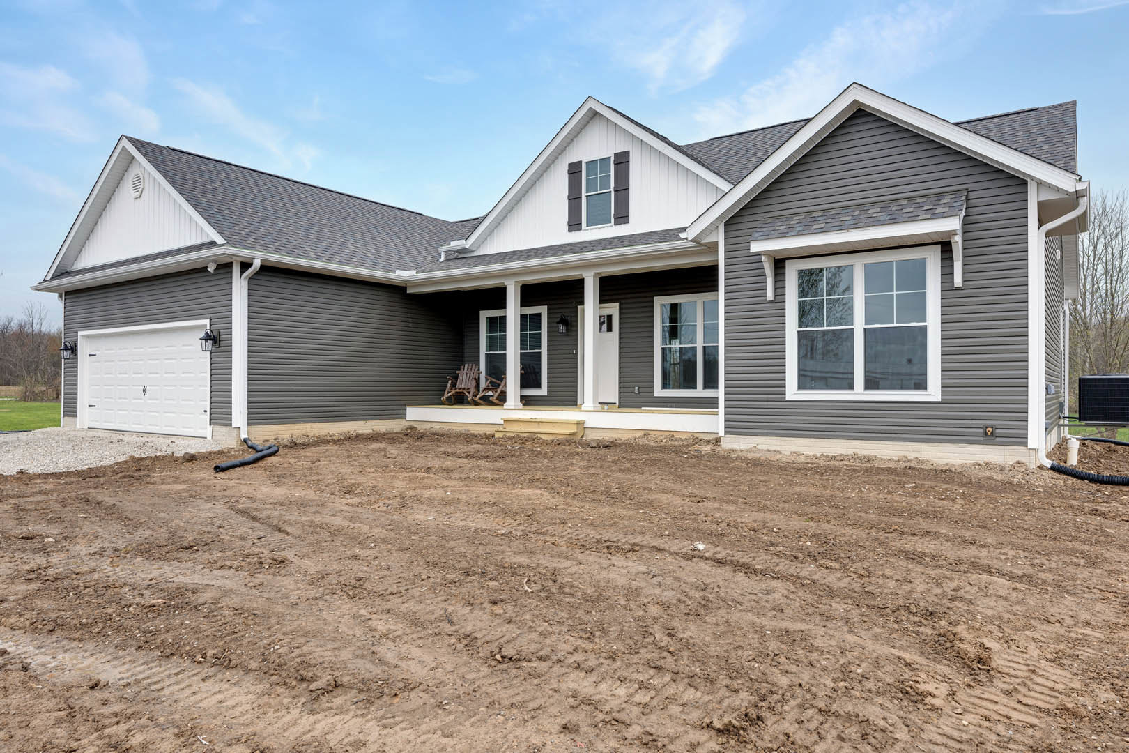 White house exterior with horizontal siding, covered front porch featuring wooden rocking chair, dirt yard in foreground, white garage with lamp post, large windows, and cloudy sky