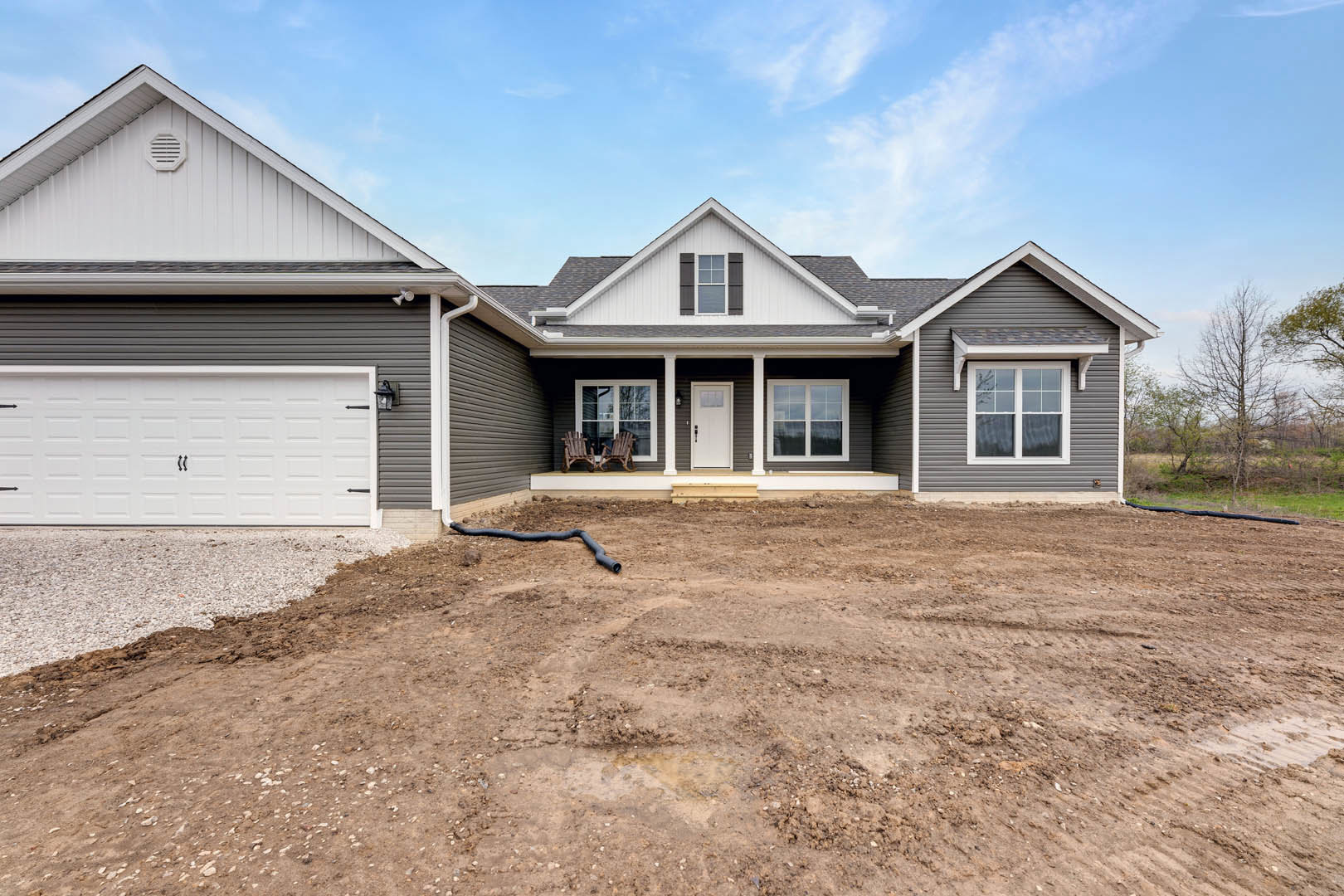 White two-story house with horizontal siding, white garage door featuring black handles, white entry door displaying a sign, white wall vent, black pipe lying on dirt yard, and