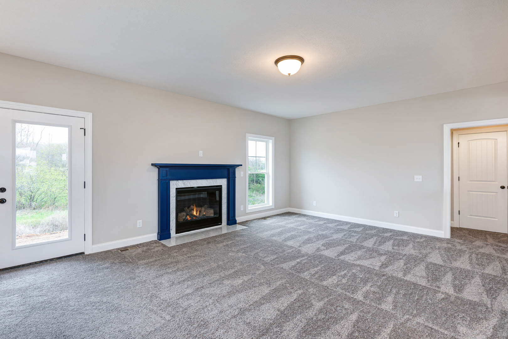Carpeted living room featuring a blue-framed fireplace, white door with black handle, ceiling light fixture, and window overlooking trees.