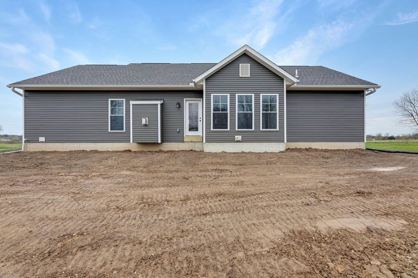 Two-story home with gray siding, white trim, glass-paneled front door, attached garage, and dirt yard with visible tire tracks