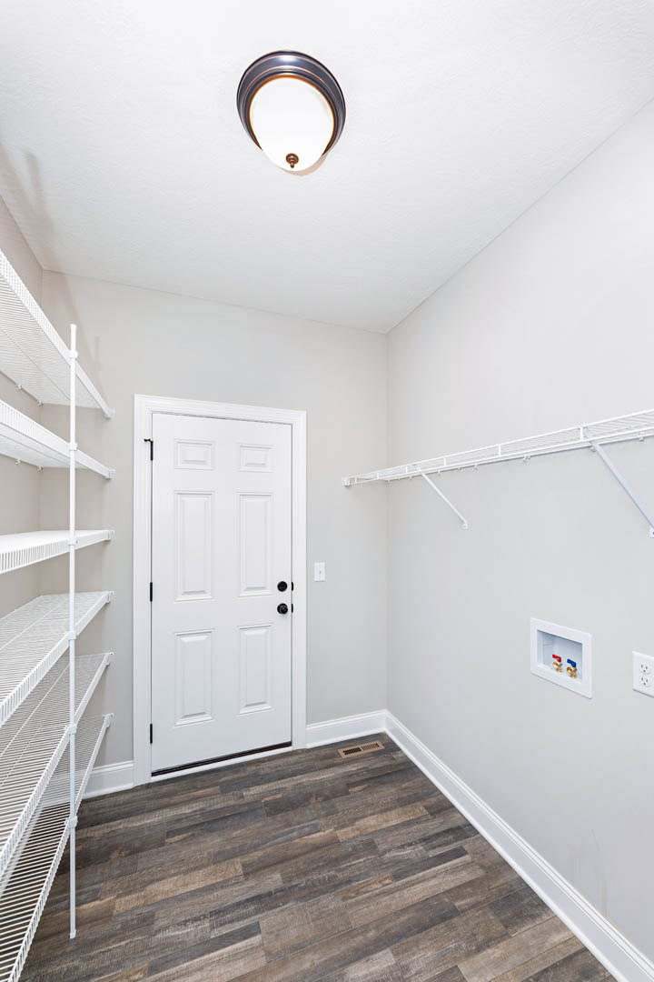 White paneled door with black hardware, wood flooring, built-in white metal shelving, ceiling light fixture, and wall-mounted box with red and blue faucet handles.