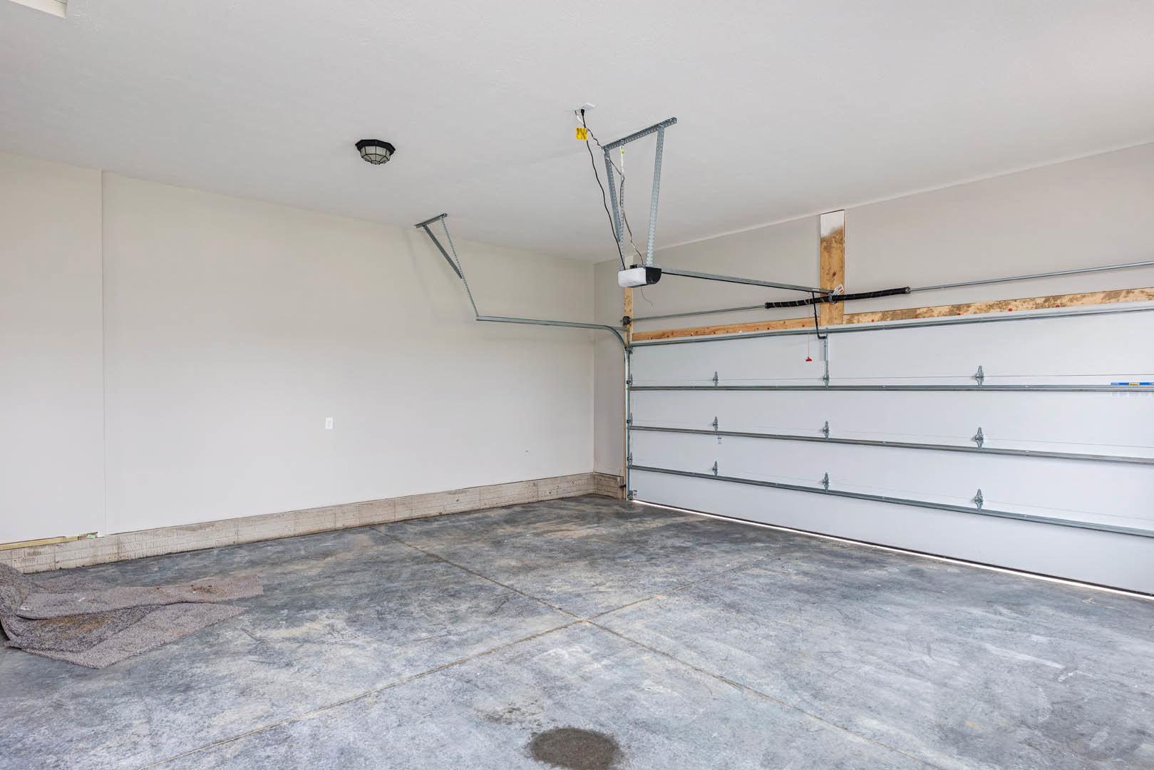 Garage interior with concrete floor featuring a central hole, metal garage door on rails, exposed metal framing and ceiling wires, plaster walls, and a circle of dirt on the