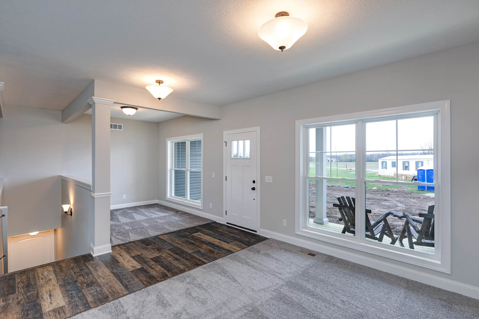 Bright room with white plaster walls, laminate wood flooring, a white door featuring a sign, large windows showing a lawn chair and trailer outside, wooden rocking chair positioned