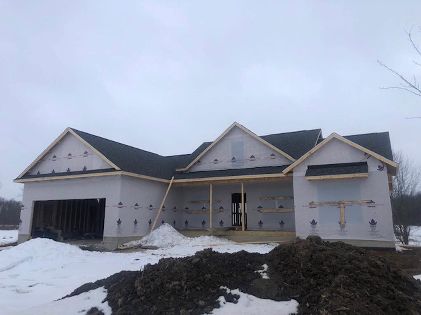Framed house under construction with exposed wood beams, black metal supports, and piles of dirt surrounded by snow