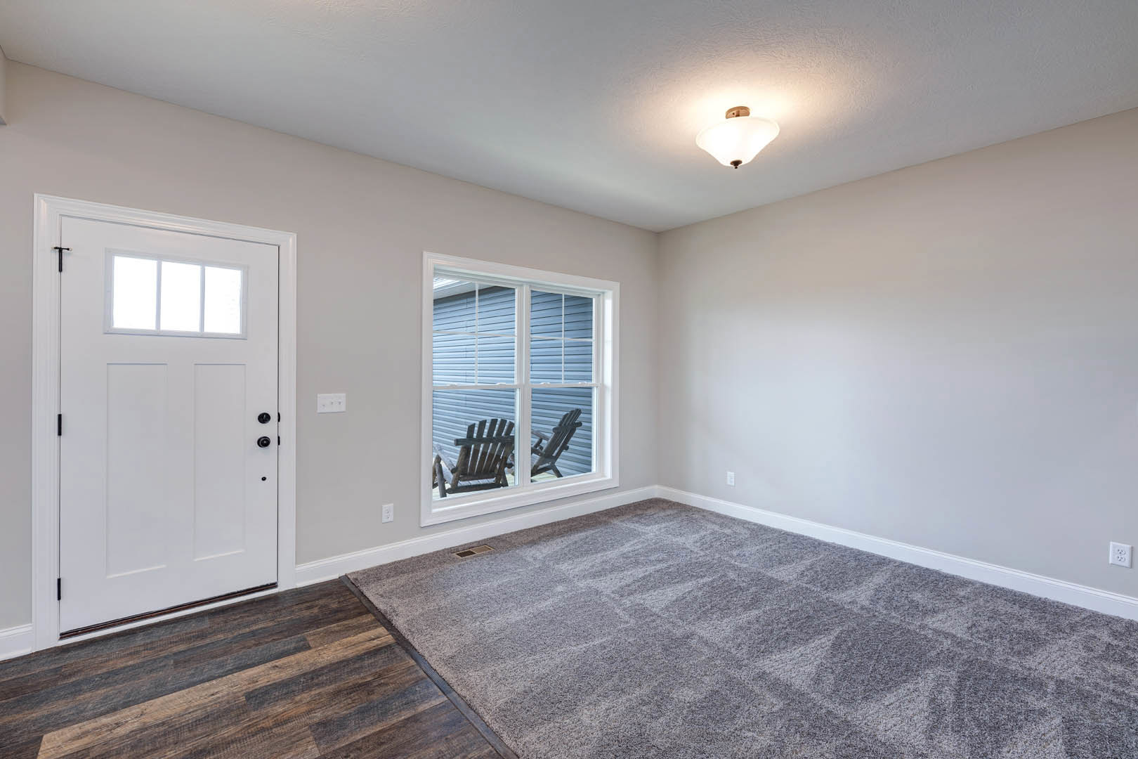 White paneled door and large window in a room with light wood laminate flooring, white plaster walls, and neutral carpet; wooden chair and small table visible outside window.
