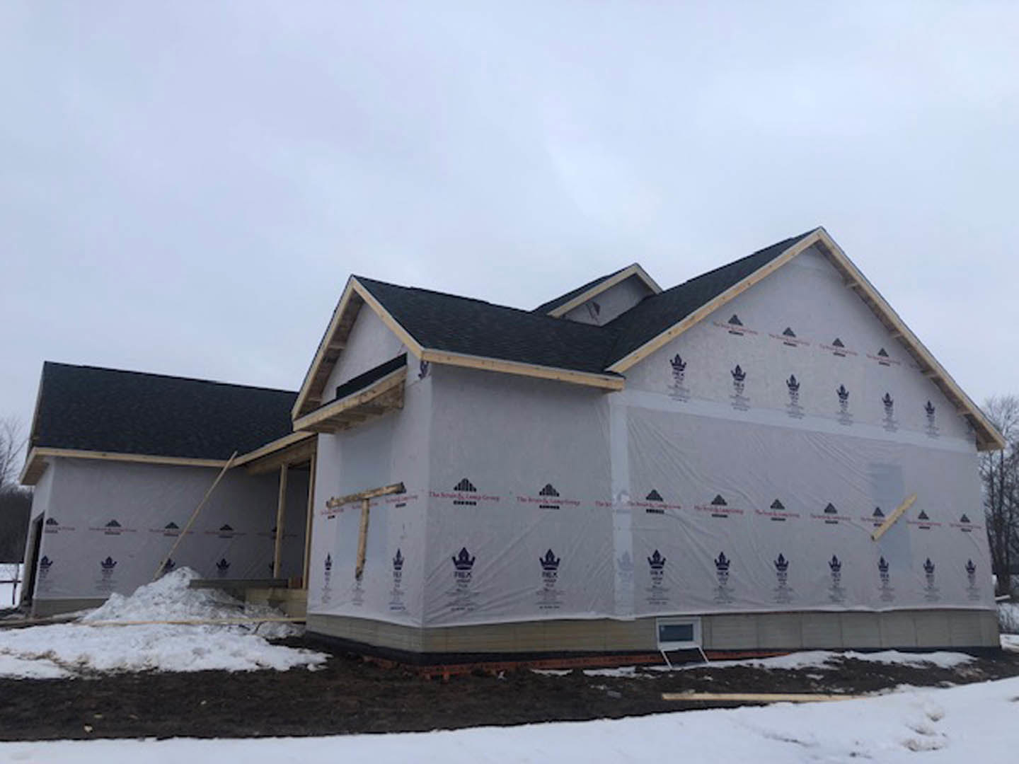 Partially built house with exposed framing and siding, snow covering the ground and roof, overcast grey sky in the background
