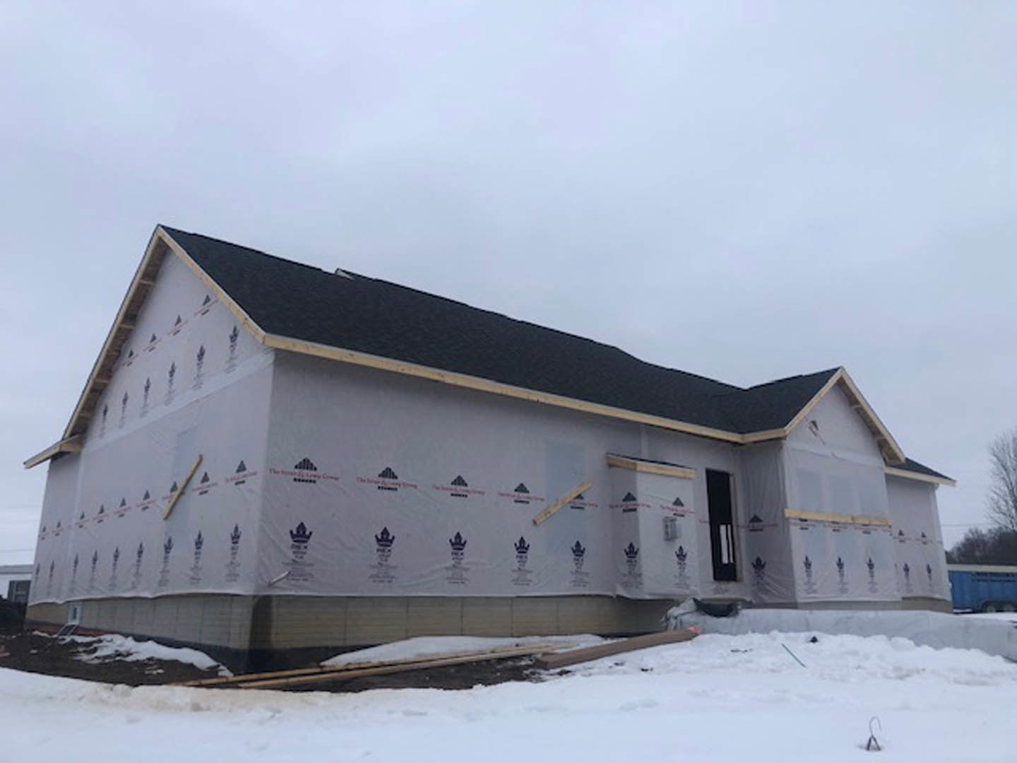 Wood-framed house under construction with exposed beams and plywood, surrounded by snow, leafless trees in background, cloudy sky overhead