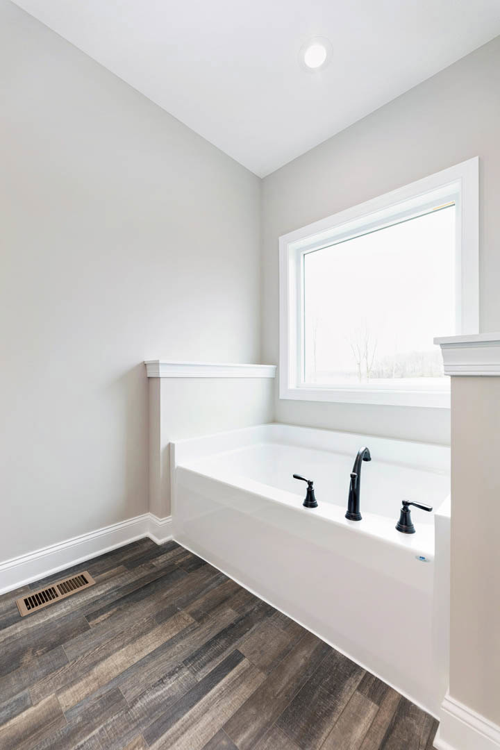 Freestanding white bathtub with matte black faucets on light wood flooring, white walls, large window with white frame, floor vent nearby