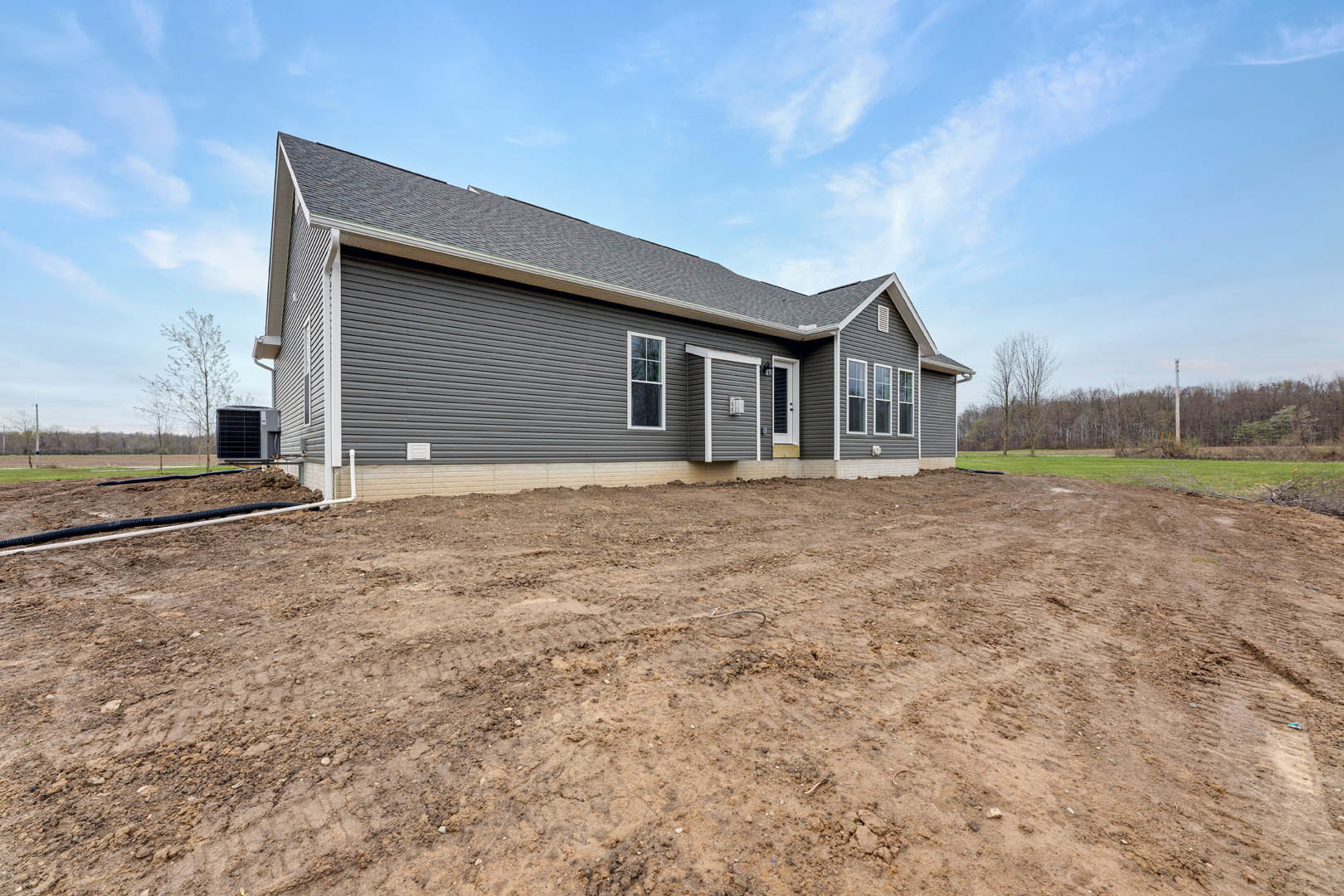 Grey custom home with white door and window frames, dirt lot in front, black utility box mounted on siding, cloudy sky overhead, sparse grass and trees in background