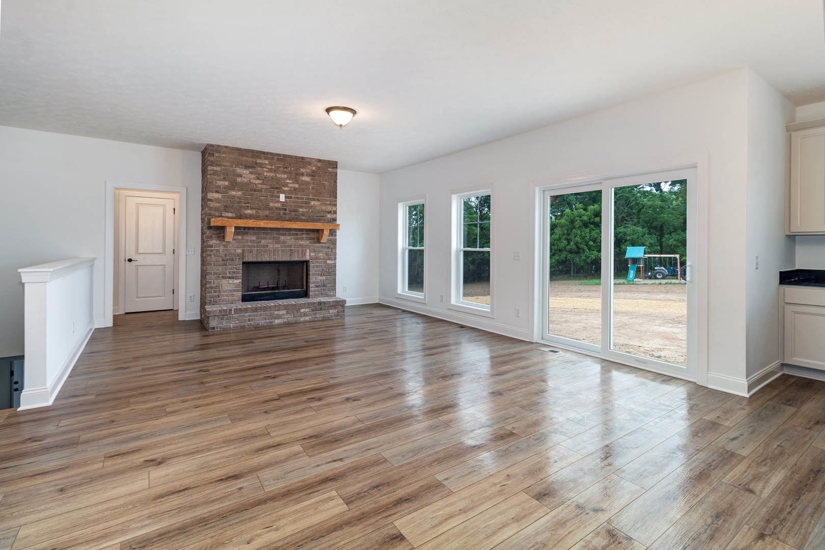 Living room with hardwood floor, white walls, central fireplace featuring a wood mantel, ceiling light fixture, and white door with black handle.