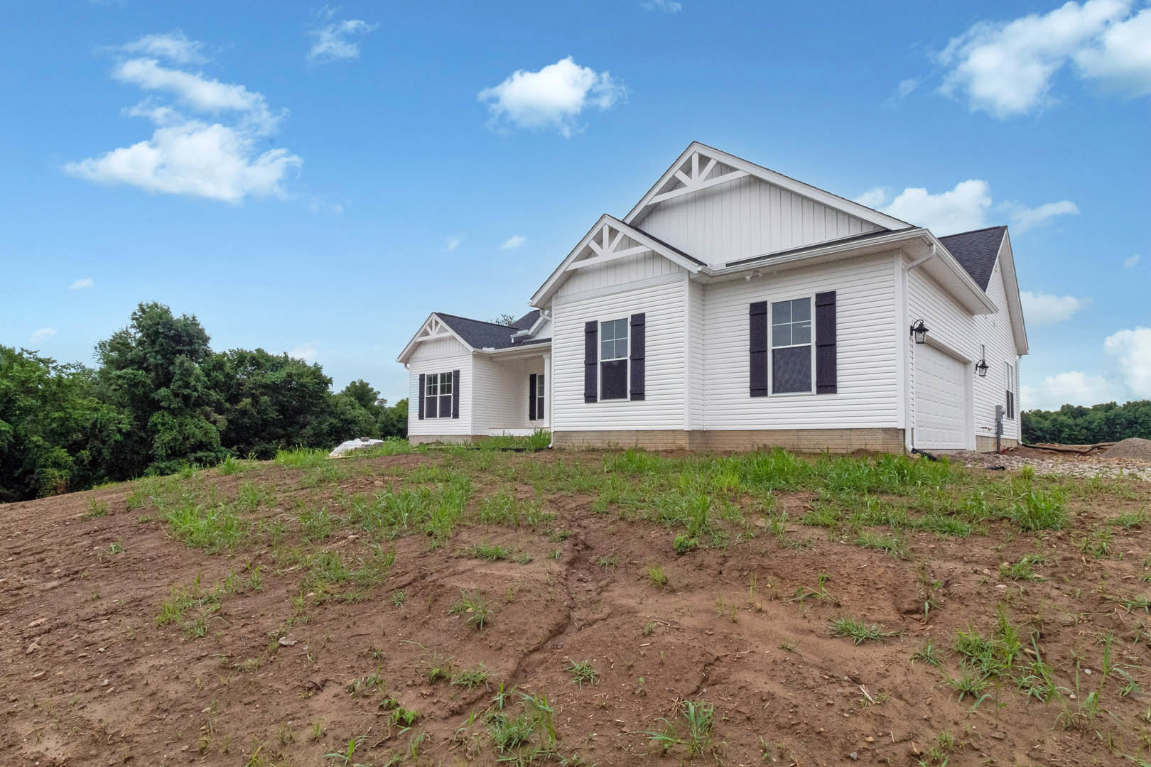 White siding house with black shutters, multi-pane windows, and white trim, set on a grassy patch with a hill and trees in the background under a cloudy sky