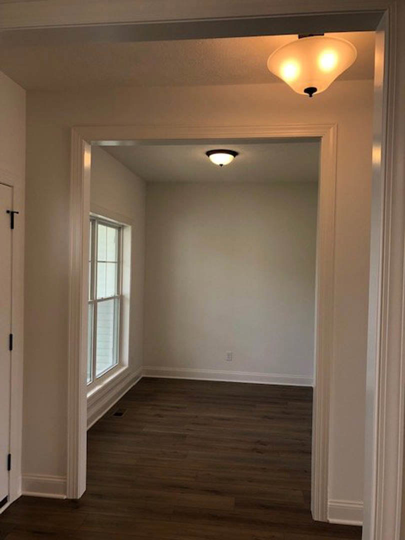 Hallway with dark wood flooring, white walls, a white-framed window, black handle on a white door, and a ceiling light fixture with two bulbs