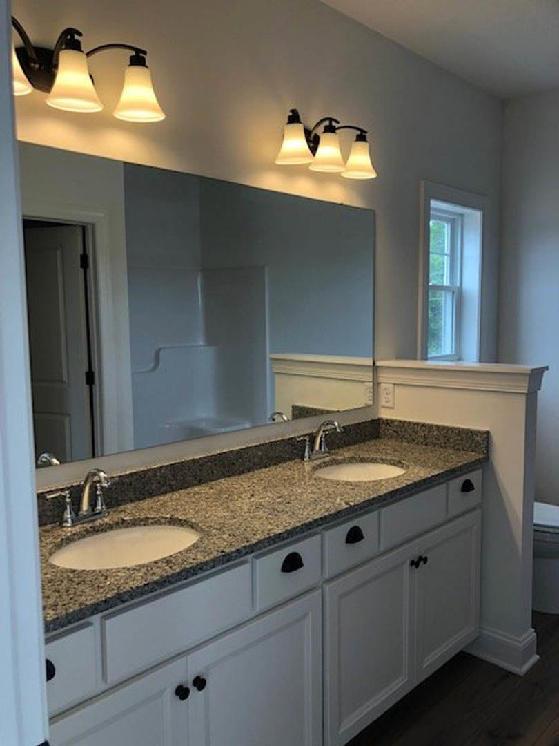 Double vanity bathroom with granite countertops, undermount sinks, large wall mirror, white cabinetry, chrome faucets, and three-light fixture above mirror; window with white frame