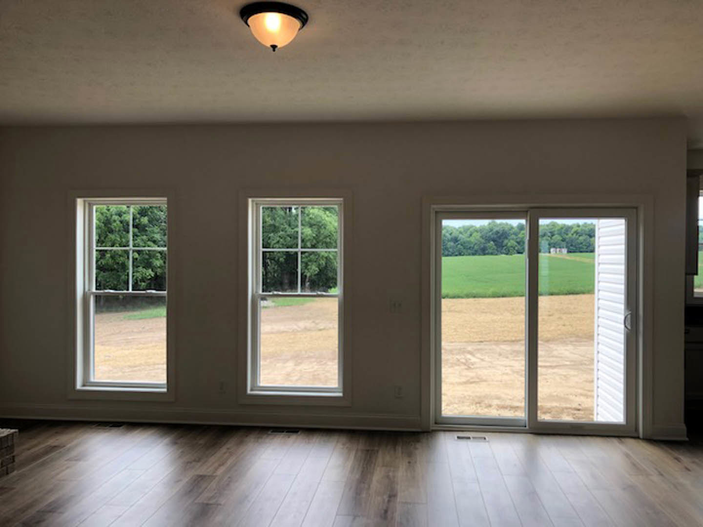 Room with wood and white tile flooring, two windows and a sliding glass door overlooking grassy field and trees, ceiling light fixture illuminated
