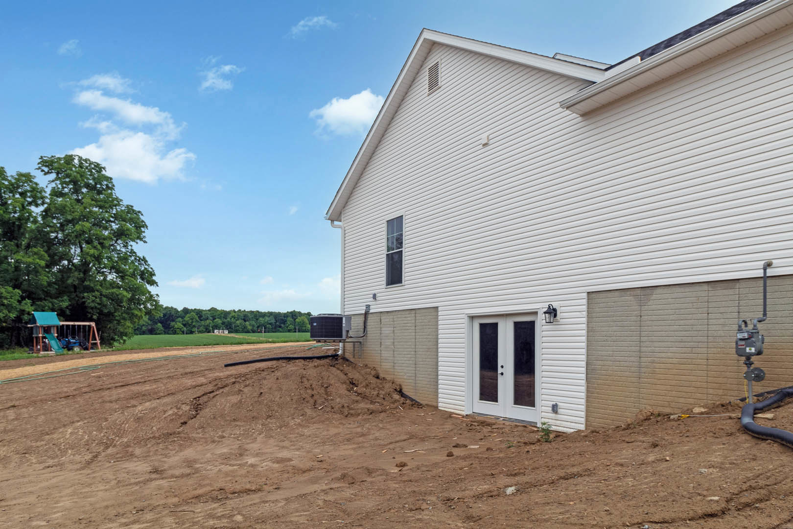 White house with glass-paneled front door, attached garage, dirt hill in foreground, mature trees and blue sky with clouds in background, large black box on pallet near driveway