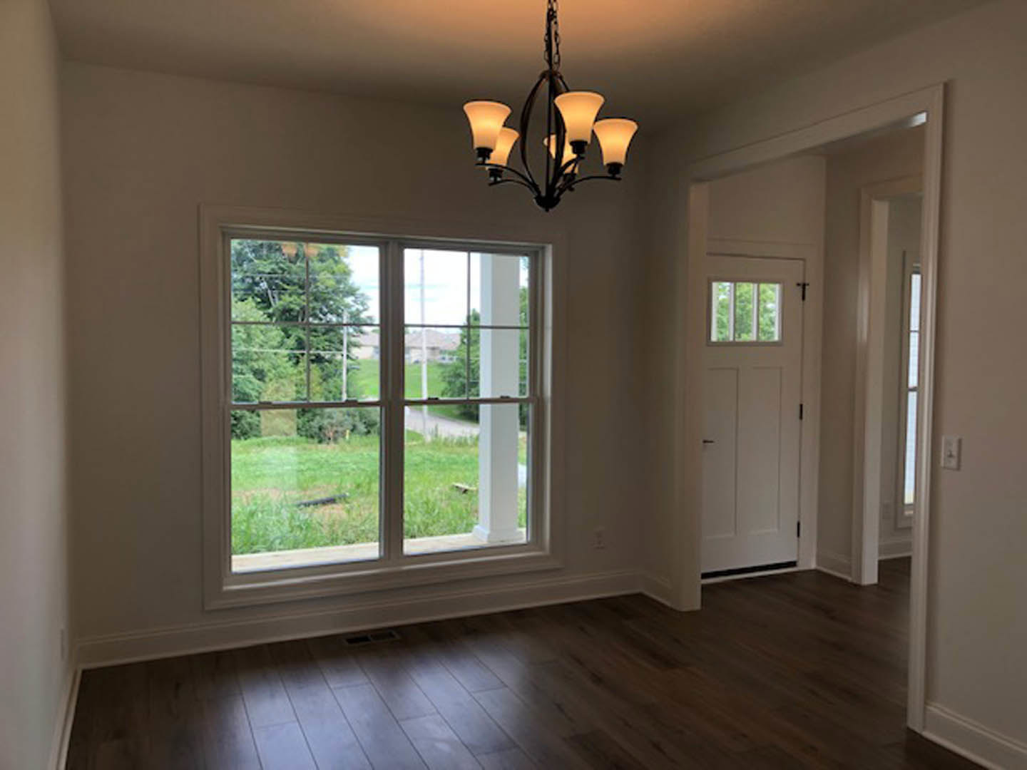 Bright room with multi-pane window overlooking grass and trees, five-light chandelier, wooden laminate flooring, white door with black handle