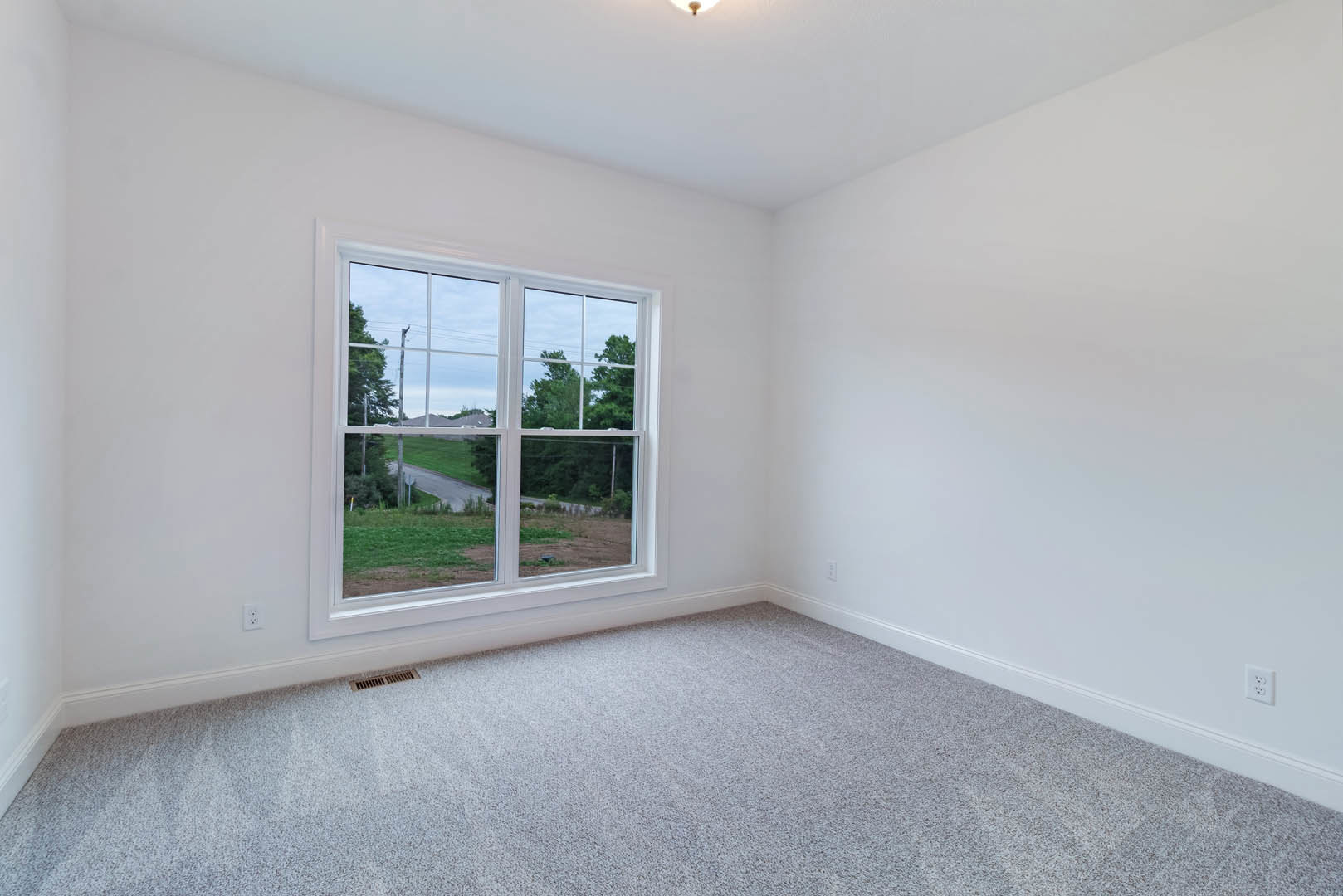 Carpeted room with white walls and large window overlooking road, grass, and trees