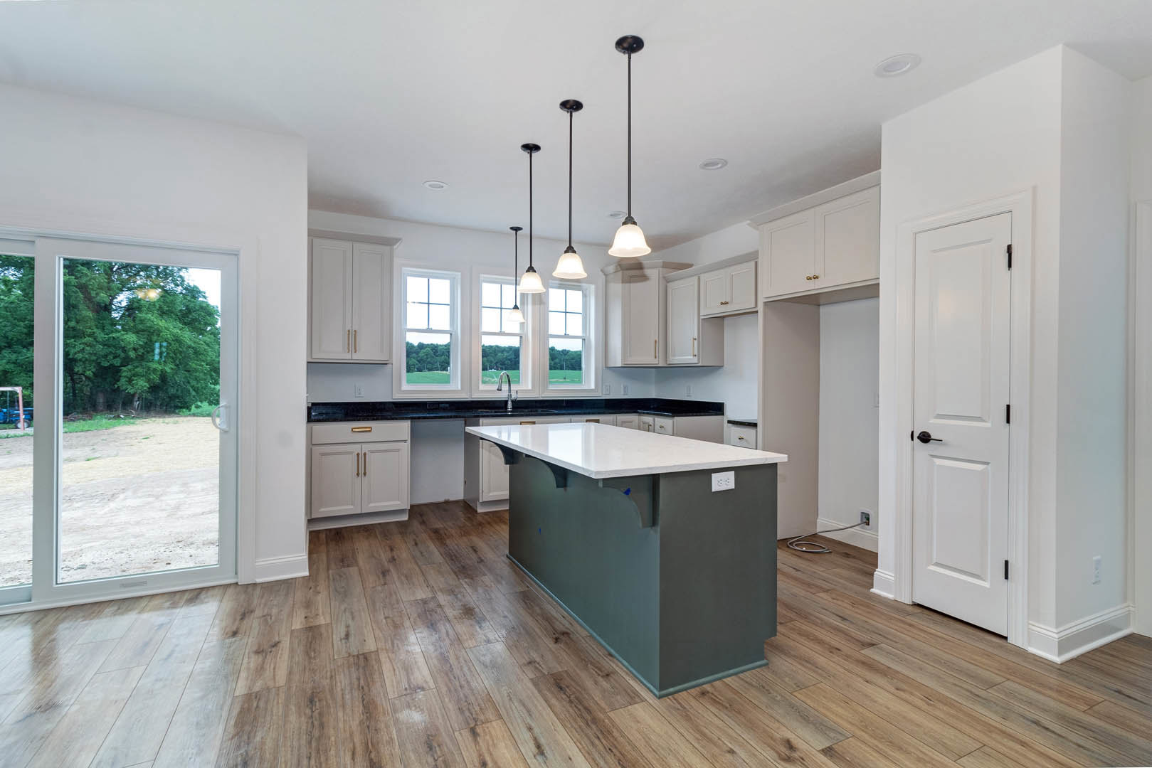 Kitchen with wood flooring, central island featuring white countertop, white cabinetry, stainless steel sink, window overlooking trees, white door with black handle, pendant light