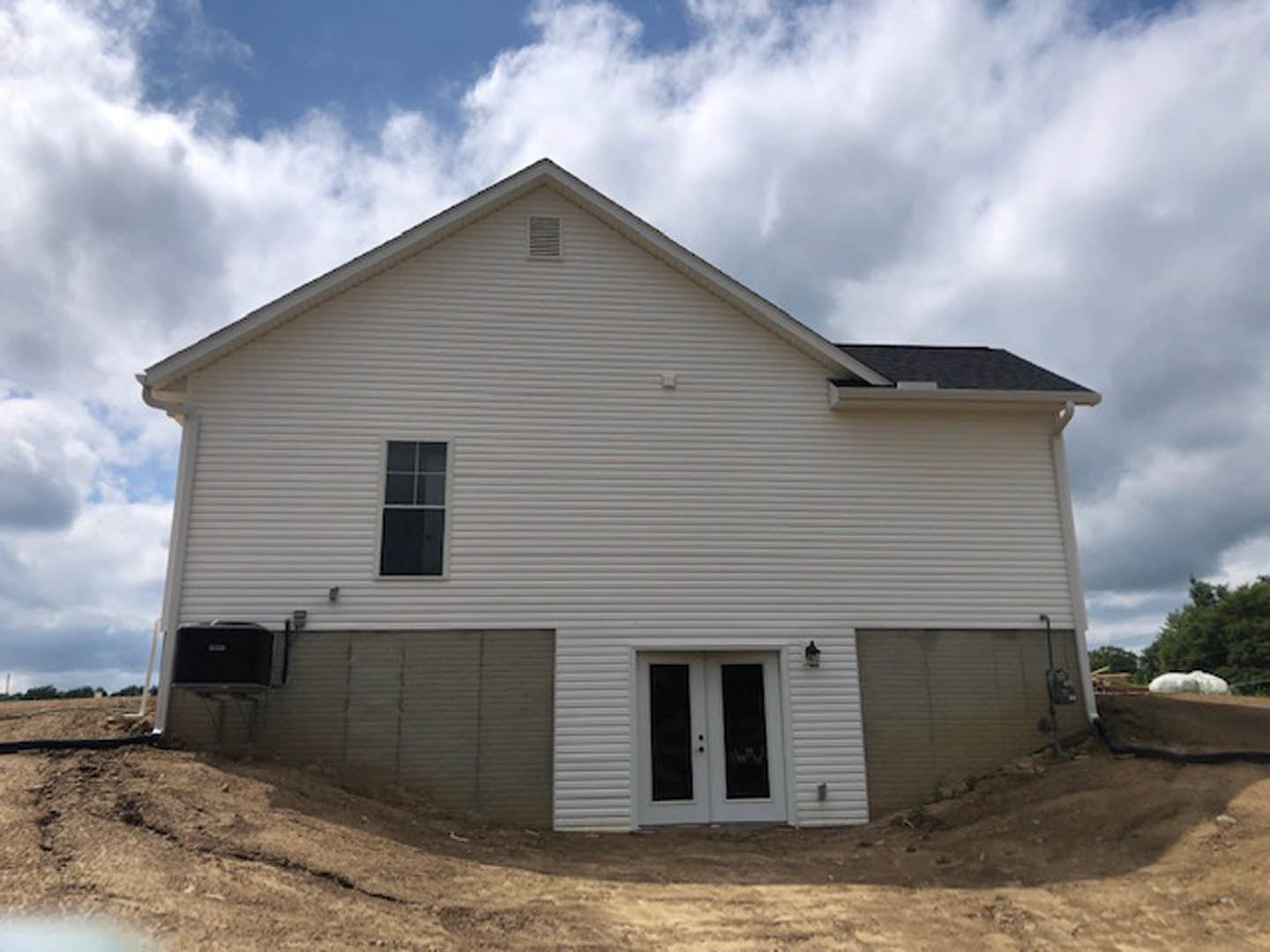 Two-story house with white siding, double glass front doors, attached garage, white shuttered window, dirt hill in foreground, cloudy sky overhead