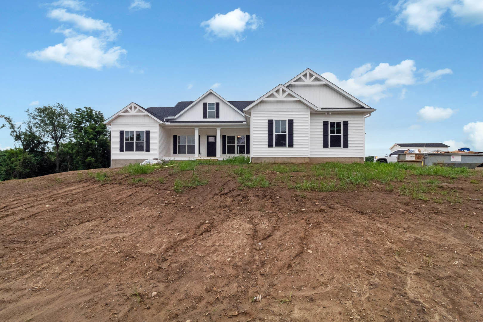 White house with black shutters and white-framed windows, set against a blue sky with clouds, surrounded by a dirt hill and grassy field, with a tree nearby.