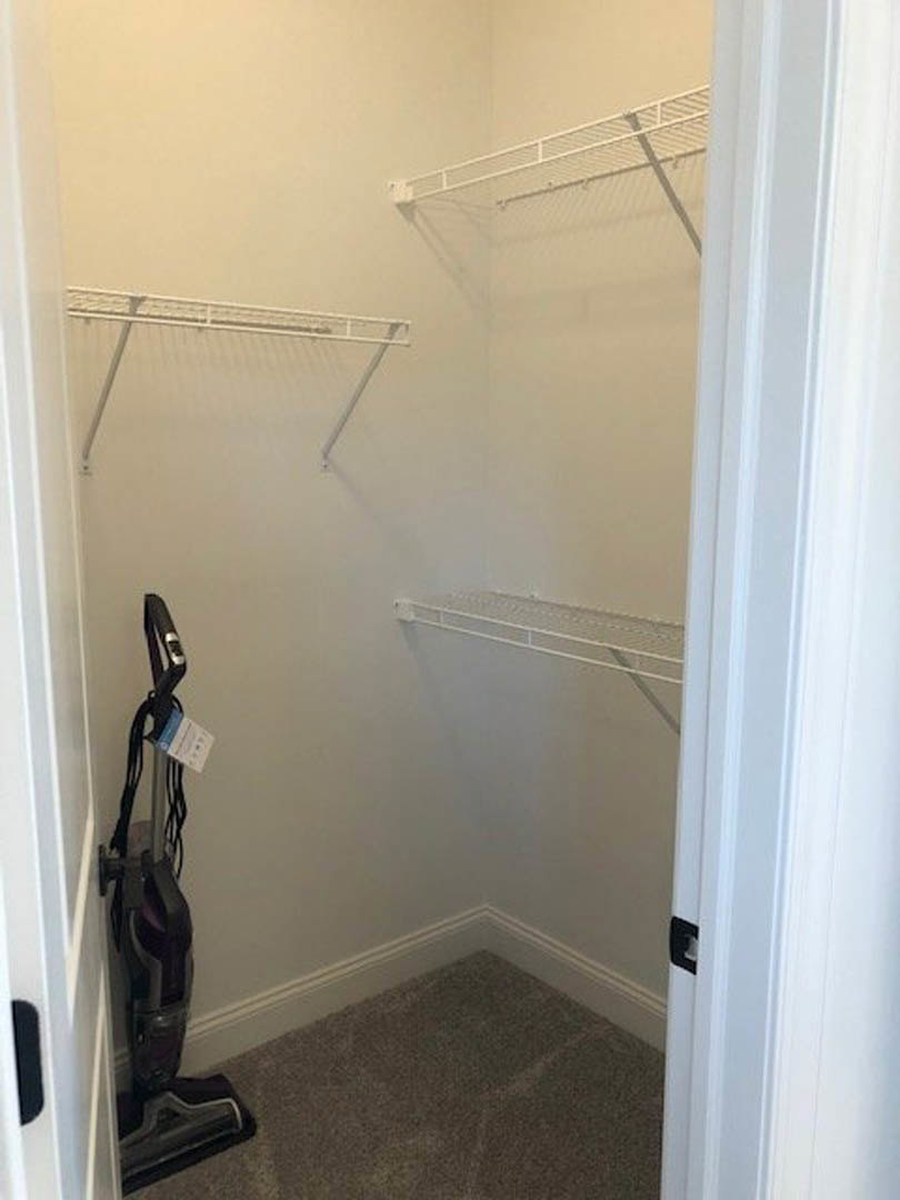 Closet with white shelves, wall-mounted vacuum cleaner, carpeted floor, white baseboard, metal bar, and white mesh panel on wall