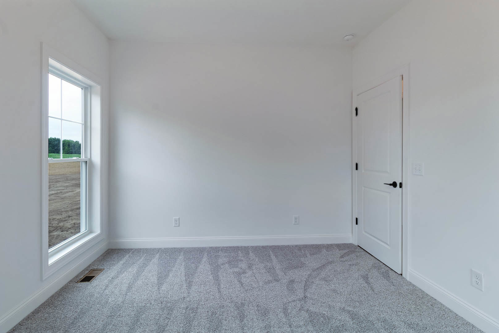 White paneled door with black handle, beige carpet flooring, large window overlooking grassy field, white walls, ceiling vent near window