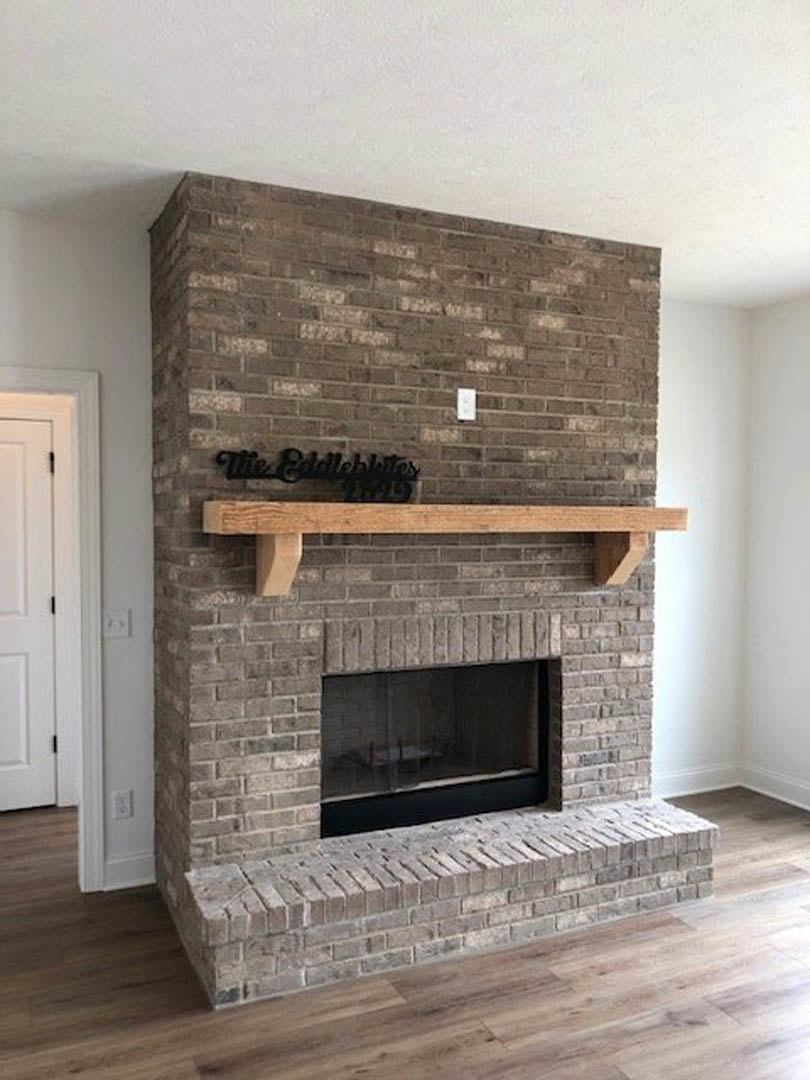 Stone fireplace with black fire screen, brick wall backdrop, wooden mantel shelf, wood flooring, large window, and decorative text on wood surface