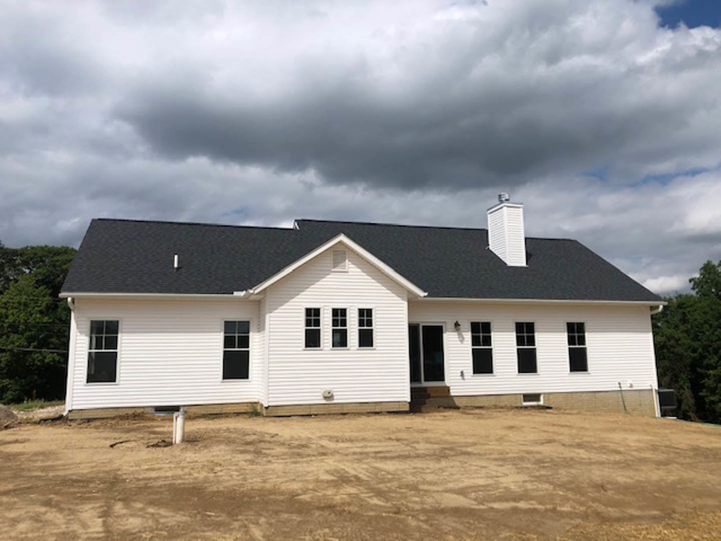 White house under construction with black roof, dirt ground in foreground, cloudy sky overhead, unfinished exterior walls, windows installed.