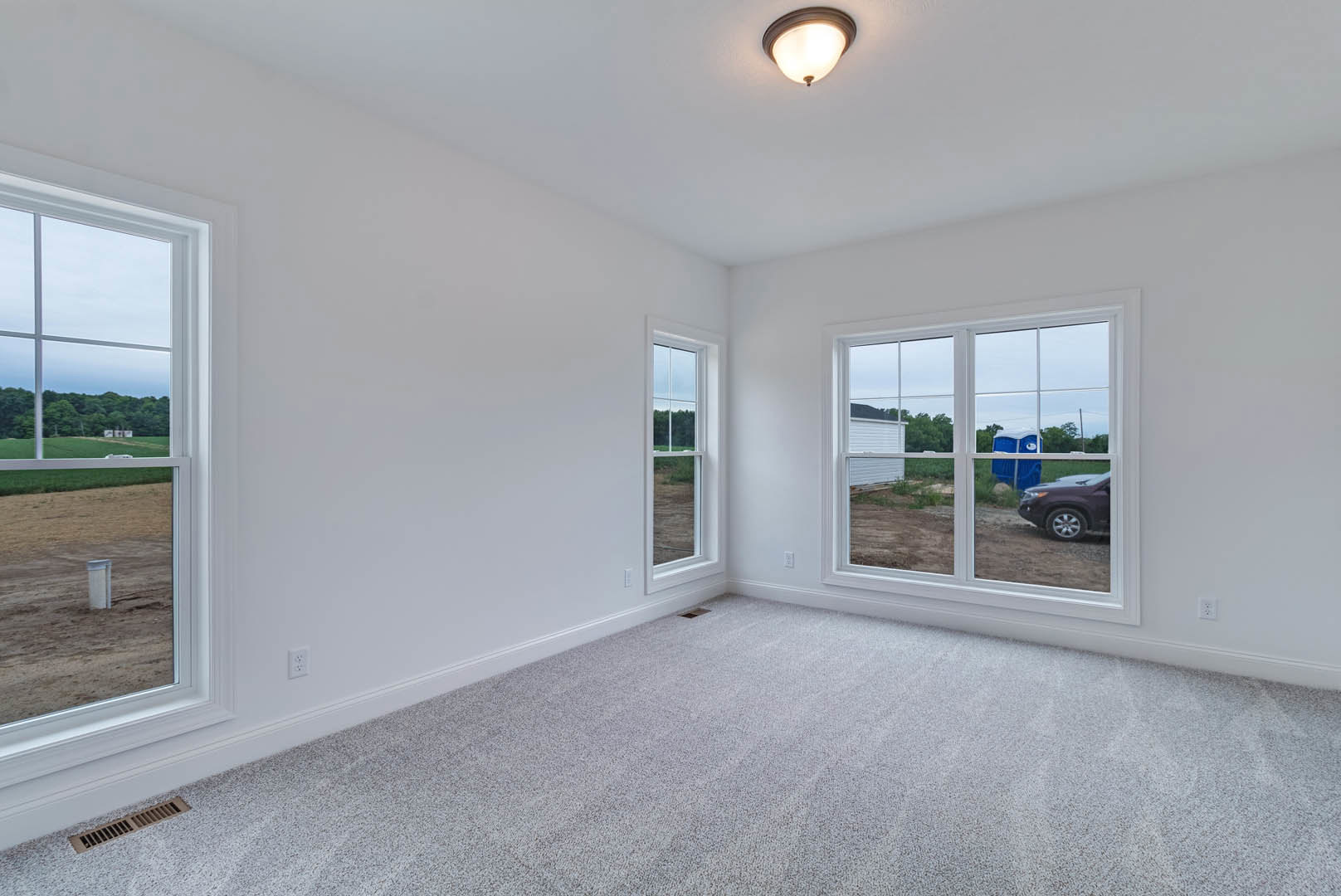 Bedroom with light-colored plaster walls, wood flooring, large window overlooking parked car, blue plastic storage bin with cartoon face, modern ceiling light fixture