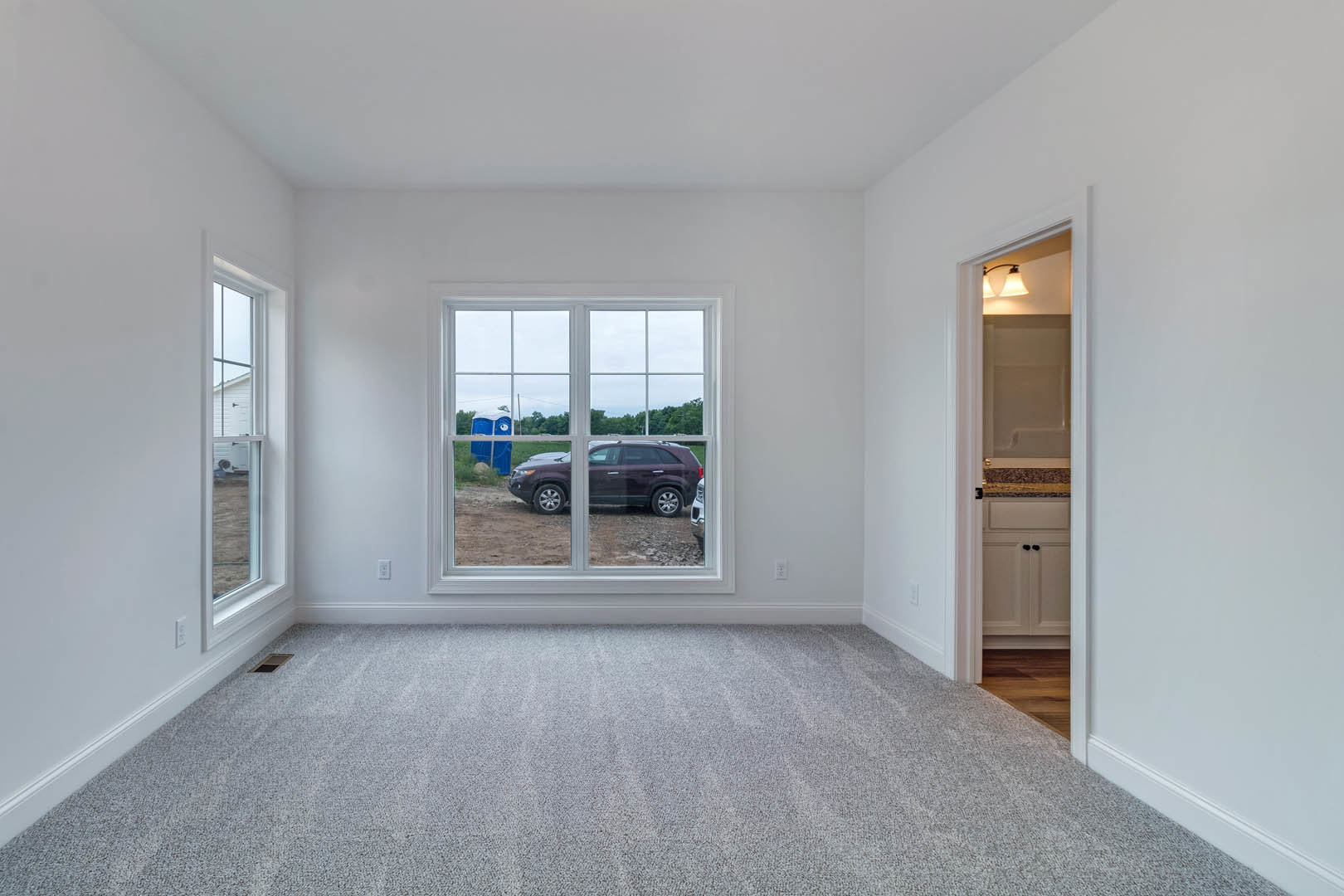 Room with laminate flooring, plaster walls, large window overlooking a parked car and portable toilet, ceiling light fixture with shade