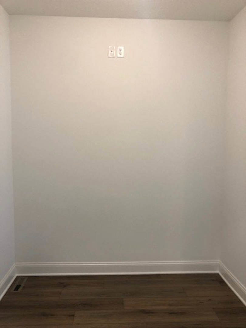White painted wall with white baseboard, wood plank flooring, visible light switch, electrical outlet, and wall vent.