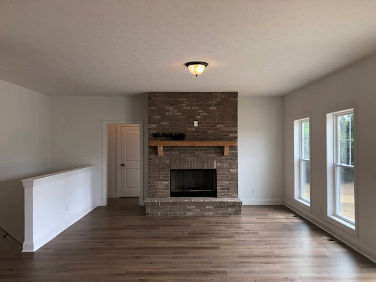 Living room with white walls, wood flooring, brick fireplace topped by a wood mantel, and a white door with a black handle.