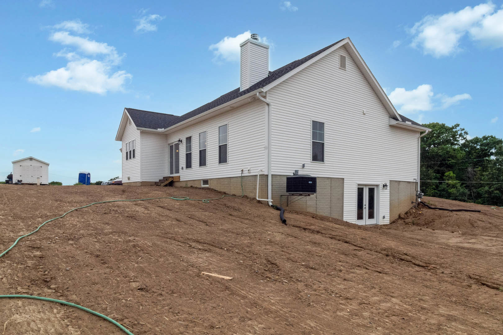 White house with a large front yard, prominent chimney, white-framed window, green hose on the lawn, and exposed pipe along the side.