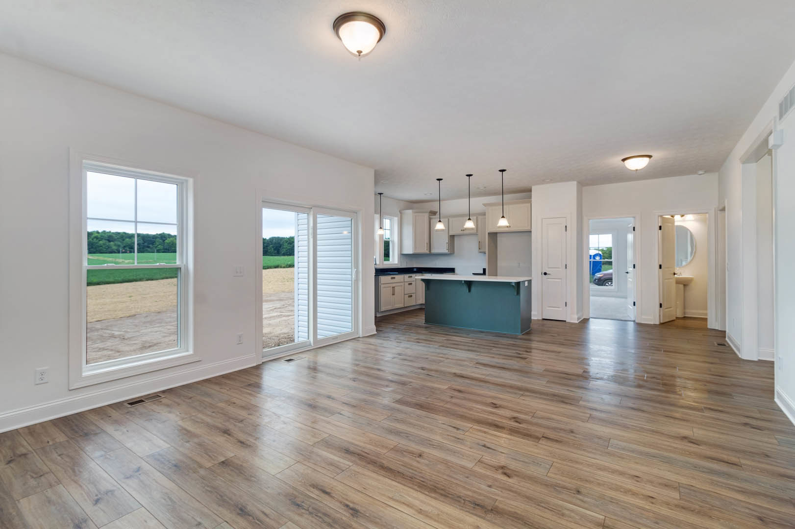Open kitchen with white cabinets and silver handles, blue accent wall, white countertop, hardwood flooring, ceiling light fixture, large window overlooking field and trees.