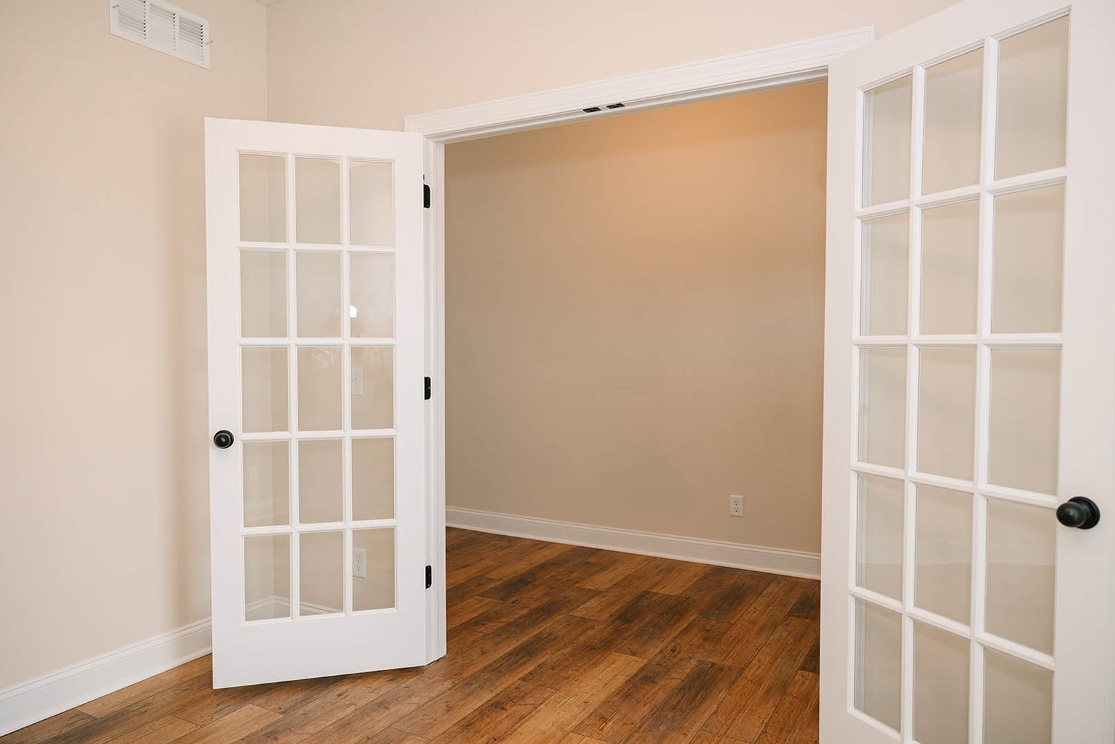 White door with glass panes open into a room with light wood flooring, white walls, and a vent visible on the wall near the door.